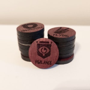 May include: Wooden game tokens with laser-etched designs. The tokens are dark brown with a red-brown top layer. One token reads "VIGILANCE" with an eye symbol, another reads "FLYING" with wings, and a third reads "INSTRUCTIBLE".
