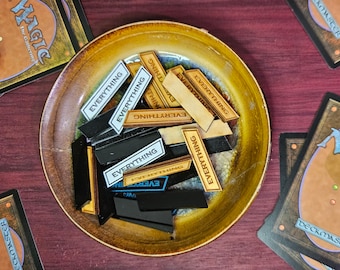 May include: A collection of rectangular game tokens with the word "EVERYTHING" printed on them, in a variety of colors, including black, white, and wood-toned. The tokens are in a gold-rimmed bowl, with Magic: The Gathering cards in the background.