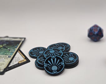 May include: A small pile of circular black tokens with a metallic blue sunburst design engraved on the surface. Beside the tokens lies a trading card with text and a multi-faceted blue and red die, all arranged on a plain white background.