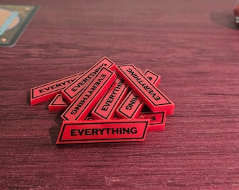 May include: A collection of red game tokens with the word "EVERYTHING" printed in black. The tokens are rectangular and stacked on a reddish-brown wooden surface. A white die and a red die are also visible, along with game cards.