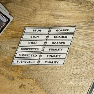 May include: A collection of white and black Magic: The Gathering game tokens with text such as "STUN", "GOADED", "SUSPECTED", and "FINALITY". The tokens are arranged on a wooden surface, along with game cards and dice.