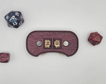 May include: A wooden dice box with two dice and a few other dice scattered around it. The box is a dark reddish-purple color with two small wooden squares in the center. The dice are various colors and sizes.