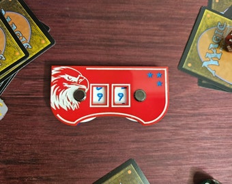May include: A red and white game counter with an eagle design and the numbers 999 displayed. The counter has three dials and is surrounded by playing cards and dice on a wooden surface.