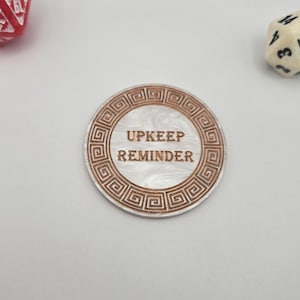May include: A round, white and brown "Upkeep Reminder" token with a Greek key border. The text is centered within the token. A red and white 20-sided die and a cream-colored 12-sided die are in the background.