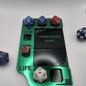 May include: A green acrylic Commander Zone dice tray with slots for dice, labeled "CASTS", "POISON", and "LIFE". The tray holds several multi-sided dice in various colors, including blue, red, and white. The text "COMMANDER ZONE" is visible.