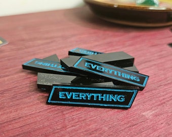 May include: A pile of black rectangular game tokens with the word "EVERYTHING" in bright blue lettering. The tokens are on a reddish-brown surface, with a few playing cards visible in the background.