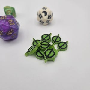 May include: A collection of green and black game tokens with a stylized design, alongside several polyhedral dice in purple, green, and off-white. The dice have visible numbers and are set against a white background.