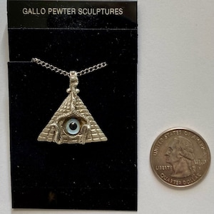 May include: A silver pewter pendant necklace featuring a pyramid with an all-seeing eye. The eye is blue with a black pupil. The pendant is hanging from a silver chain. The pendant is displayed on a black background with a quarter for size comparison. The card behind the pendant reads "Gallo Pewter Sculptures".