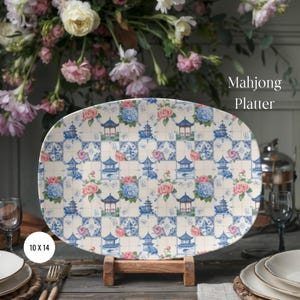 May include: An oval ceramic Mahjong Platter measuring 10 by 14 inches. The surface features a repeating grid pattern of blue pagodas, pink and blue floral clusters, and small Mahjong tile icons on a cream background, displayed on a wooden stand.