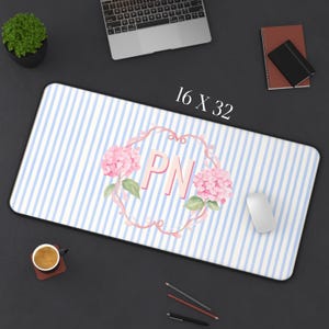 May include: A rectangular desk mat featuring a light blue and white vertical striped pattern. In the center, a pink floral wreath surrounds the letters PN. The text 16 X 32 is printed above the mat, which is displayed on a dark surface.