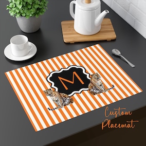 May include: Custom placemat with orange and white stripes, featuring a black and white emblem with the letter "M" and two leopard illustrations. The text "Custom Placemat" is in the lower right corner.