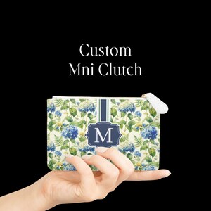 May include: A rectangular mini clutch featuring a floral pattern of blue hydrangeas and green leaves on a cream background. A central vertical navy blue stripe contains a dark blue emblem with the white letter M. Text reads Custom Mni Clutch.