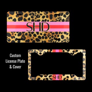 May include: A rectangular license plate and a matching license plate frame, both featuring a brown and black leopard print pattern. A horizontal pink and red stripe runs across the center of each, with the black letters SHD printed on the plate.