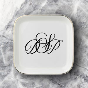 May include: A square white ceramic trinket dish with rounded corners and a thin gold-colored metallic rim. The center of the dish features a black decorative monogram with the stylized cursive letters D, S, and P intertwined in a single design.