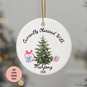 May include: A round white ceramic ornament hanging from a gold string. The design features a green Christmas tree, pink gift ribbon, and mahjong tiles. Black text reads Currently Obsessed With Mahjong and the year 2026 is printed at the bottom.