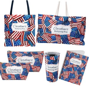 May include: A collection of patriotic-themed accessories featuring a pattern of American flags. Items include tote bags, cosmetic bags, a travel mug, and a spiral-bound notebook. Each item has a white label with the text "Caroline's Mahjong Bag" or "Caroline's Mahjong Journal".