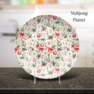 May include: A round ceramic platter featuring a festive pattern of Mahjong tiles, red and green Christmas ornaments, pine branches, holly berries, and red bows. The text Mahjong Platter is printed in the upper right corner of the image.