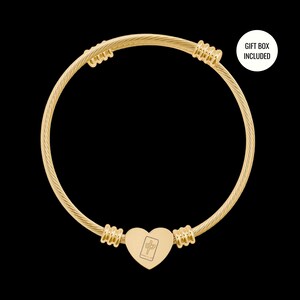May include: A gold-toned cable wire bangle bracelet featuring a central heart-shaped charm engraved with a mahjong tile symbol. The bracelet includes ribbed metal accents on either side of the heart charm and a white circular label stating GIFT BOX INCLUDED.