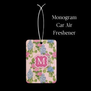 May include: A rectangular car air freshener with a white hanging string, featuring a pink background with blue and white ginger jars, green leaves, and pink hydrangea flowers. A central pink scalloped square contains the white letter M. Text reads Monogram Car Air Freshener.