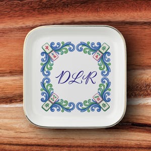 May include: A square white ceramic trinket dish with rounded corners and a gold-toned rim. The center features the blue monogram DLR surrounded by a decorative border of blue and green swirls interspersed with four colorful mahjong tiles.
