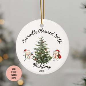 May include: A round white ceramic Christmas ornament hanging from a gold string. It features a central green Christmas tree, two cartoon Mahjong tiles wearing Santa hats, and the black text Currently Obsessed With Mahjong 2026.