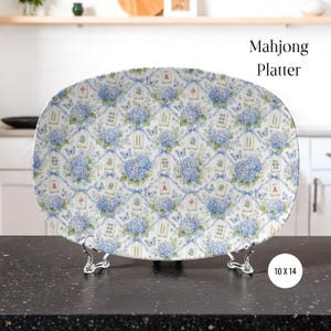 May include: An oval platter featuring a repeating pattern of blue hydrangea flowers, light blue ribbons, butterflies, and Mahjong tiles. The text "Mahjong Platter" and "10 X 14" are visible. The platter is displayed on a silver-toned metal stand.