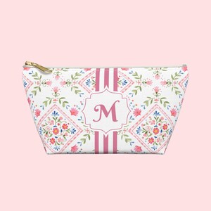 Puede incluir: Un neceser blanco con un estampado floral en rosa, azul y verde. La bolsa presenta una inicial "M" rosa en un marco decorativo, con rayas rosas y una cremallera dorada.