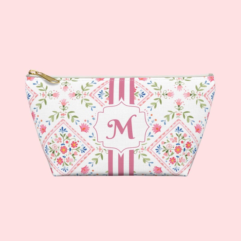 Puede incluir: Un neceser blanco con un estampado floral en rosa, azul y verde. La bolsa presenta una inicial "M" rosa en un marco decorativo, con rayas rosas y una cremallera dorada.