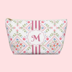 Puede incluir: Un neceser blanco con un estampado floral en rosa, azul y verde. La bolsa presenta una inicial "M" rosa dentro de un marco decorativo, con rayas rosas en la parte delantera. Una cremallera dorada asegura la parte superior.