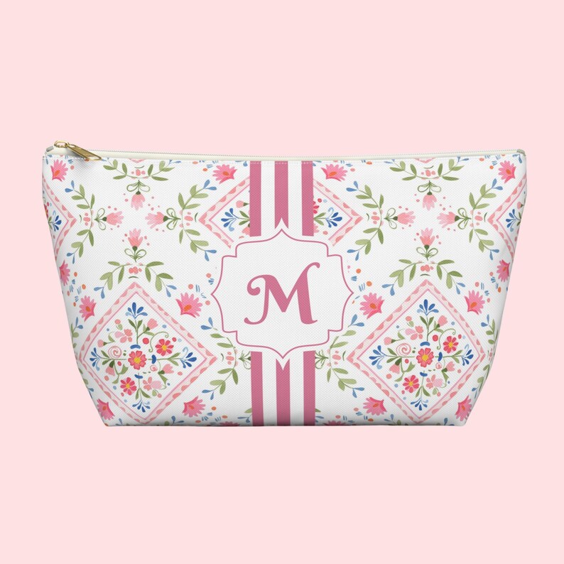 Puede incluir: Un neceser blanco con un estampado floral en rosa, azul y verde. La bolsa presenta una inicial "M" rosa dentro de un marco decorativo, con rayas rosas en la parte delantera. Una cremallera dorada asegura la parte superior.