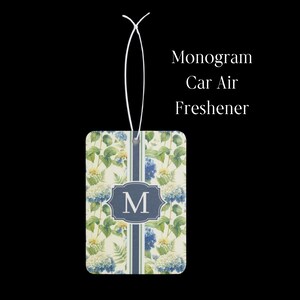 May include: A rectangular car air freshener hanging from a white string. The surface features a floral pattern with blue and white hydrangea blossoms and green leaves. A central blue vertical stripe contains a dark blue frame with the letter M.