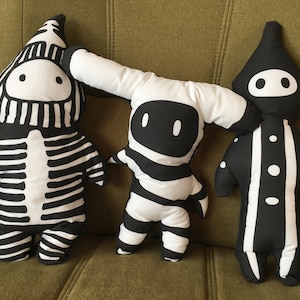 May include: Three black and white striped plush toys. The first toy is a skeleton with a hood. The second toy is a robot with a round head. The third toy is a hooded figure with a circle pattern.