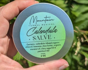 May include: A silver tin of calendula salve with a green and blue label. The label reads "Mountain Herbal Revival Calendula Salve" and lists the ingredients: calendula infused organic olive oil, beeswax, shea butter, argan oil, clary sage EO & patchouli EO. The tin is labeled 2 oz.