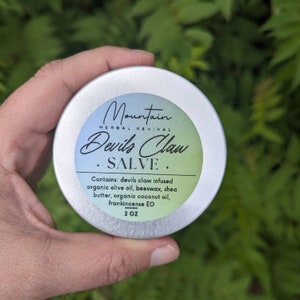 May include: A silver tin with a black label that reads "Mountain Herbal Revival Devils Claw + SALVE +" and "Contains: devils claw infused organic olive oil, beeswax, shea butter, organic coconut oil, frankincense EO 2 OZ".