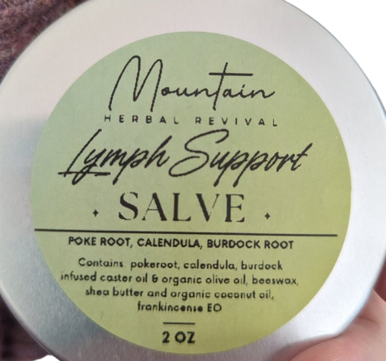 May include: A silver tin with a green label that reads "Mountain Herbal Revival Lymph Support Salve". The label also lists the ingredients: "Poke Root, Calendula, Burdock Root". It also says "Contains pokeroot, calendula, burdock infused caster oil & organic olive oil, beeswax, shea butter and organic coconut oil, frankincense EO". The label also says "2 OZ".