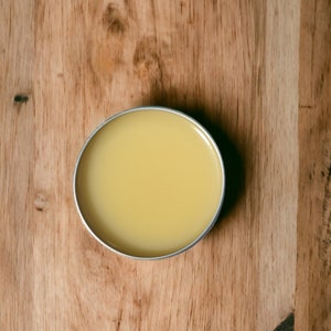 May include: A round metal tin filled with a light yellow balm or salve. The tin is sitting on a wooden surface.