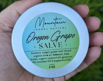 May include: A silver tin with a green and white label that reads "Mountain Herbal Revival Oregon Grape • SALVE •" and lists the ingredients: "oregon grape root infused organic olive oil, beeswax, shea butter, organic coconut oil, geranium EO, lavender EO & peppermint EO". The tin is labeled "2 OZ".