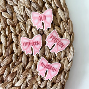 May include: Four pink bow stickers with names written on them. The names are Ava, Kathleen, Kristin, and Elizabeth.