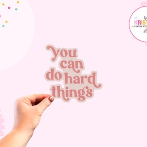 May include: A sticker with the text "You can do hard things" in a retro font. The sticker is held by a hand against a light pink background. The sticker has a light pink and beige color scheme.