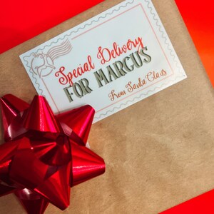 K&ouml;nnte beinhalten: Eine braune Papiert&uuml;te mit einer roten Schleife und einem wei&szlig;en Etikett mit der Aufschrift "Special Delivery for Marcus From Santa Claus".