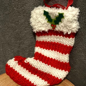 May include: Red and white striped knitted Christmas stocking with a white fluffy cuff and a green holly appliqué with red berries. The stocking has a red ribbon loop for hanging.