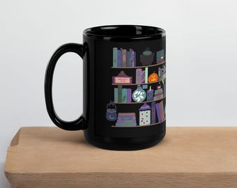 May include: Black ceramic mug featuring a whimsical illustration of a bookshelf filled with potion bottles, books, and a cauldron. The design includes a pumpkin, candles, and a crescent moon. The mug is placed on a wooden surface.