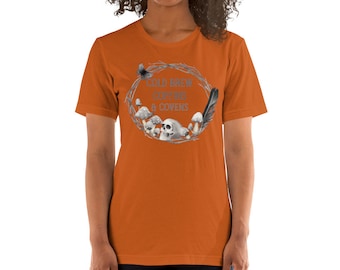 Może przedstawiać: Pomarańczowy T-shirt z krótkim rękawem z grafiką przedstawiającą wieniec z gałęzi, grzybów, czaszki, pióra i motyla. Tekst na koszulce brzmi "Cold Brew Coffee & Covens".