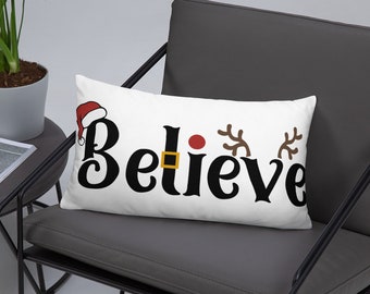May include: White throw pillow with the word "Believe" in black cursive lettering. A red Santa hat, a red nose, and two reindeer antlers are incorporated into the design.
