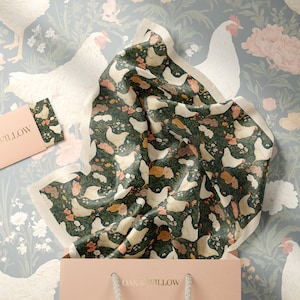 May include: A gift bag with a floral patterned scarf featuring chickens, flowers, and foliage. The scarf is dark green with white, orange, and brown accents. A matching card and a floral wallpaper background complete the scene. The gift bag has the text "OAK & WILLOW".