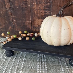 May include: A black wooden tiered stand with rounded legs, featuring a white pumpkin with red speckles on top. The stand is on a green and white patterned surface.
