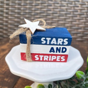 Puede incluir: Una pila de tres bloques de madera pintados en rojo, blanco y azul con las palabras "Stars and Stripes" pintadas en ellos. Una estrella de madera blanca está en la parte superior de los bloques.