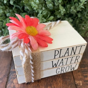 May include: Three white wooden blocks stacked on top of each other. The top block says "PLANT", the middle block says "WATER", and the bottom block says "GROW". A pink flower with a brown twine bow is tied around the blocks.