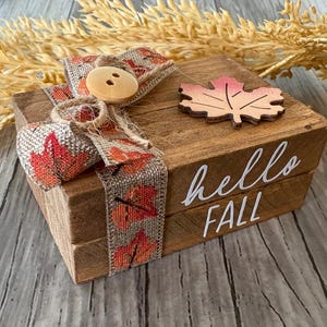May include: A stack of two small wooden blocks with a natural brown finish, featuring the white text "hello FALL" on the side. The blocks are wrapped in a burlap ribbon with a red and orange leaf pattern, a wooden button, and a wooden leaf cutout.