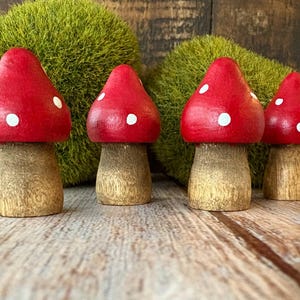 May include: Four wooden mushroom figurines with red caps and white spots. The mushroom stems are a light brown color. The mushrooms are in front of a green mossy background, and are sitting on a wooden surface.